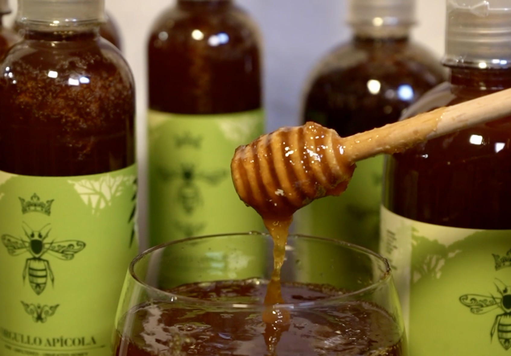A wooden dipper dripping raw honey into a glass, with green Orgullo Apícola bottles behind it